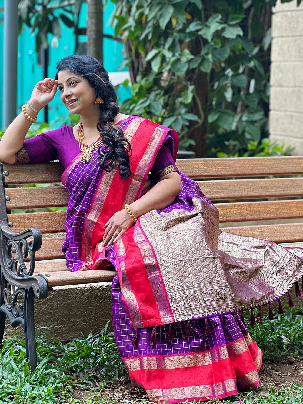 Woman sitting on a bench in purple silk saree with red border.
