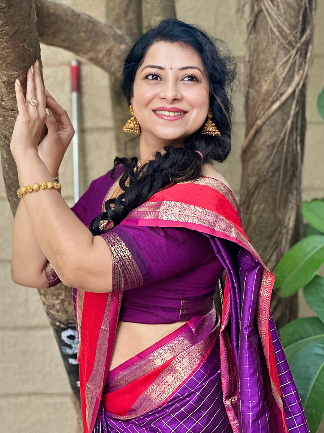 Woman in purple silk saree with red border, smiling by a tree.