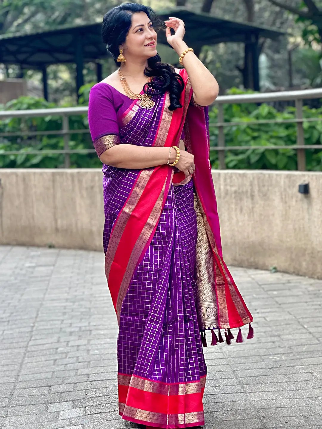 Woman wearing purple silk saree with red border, standing outdoors.