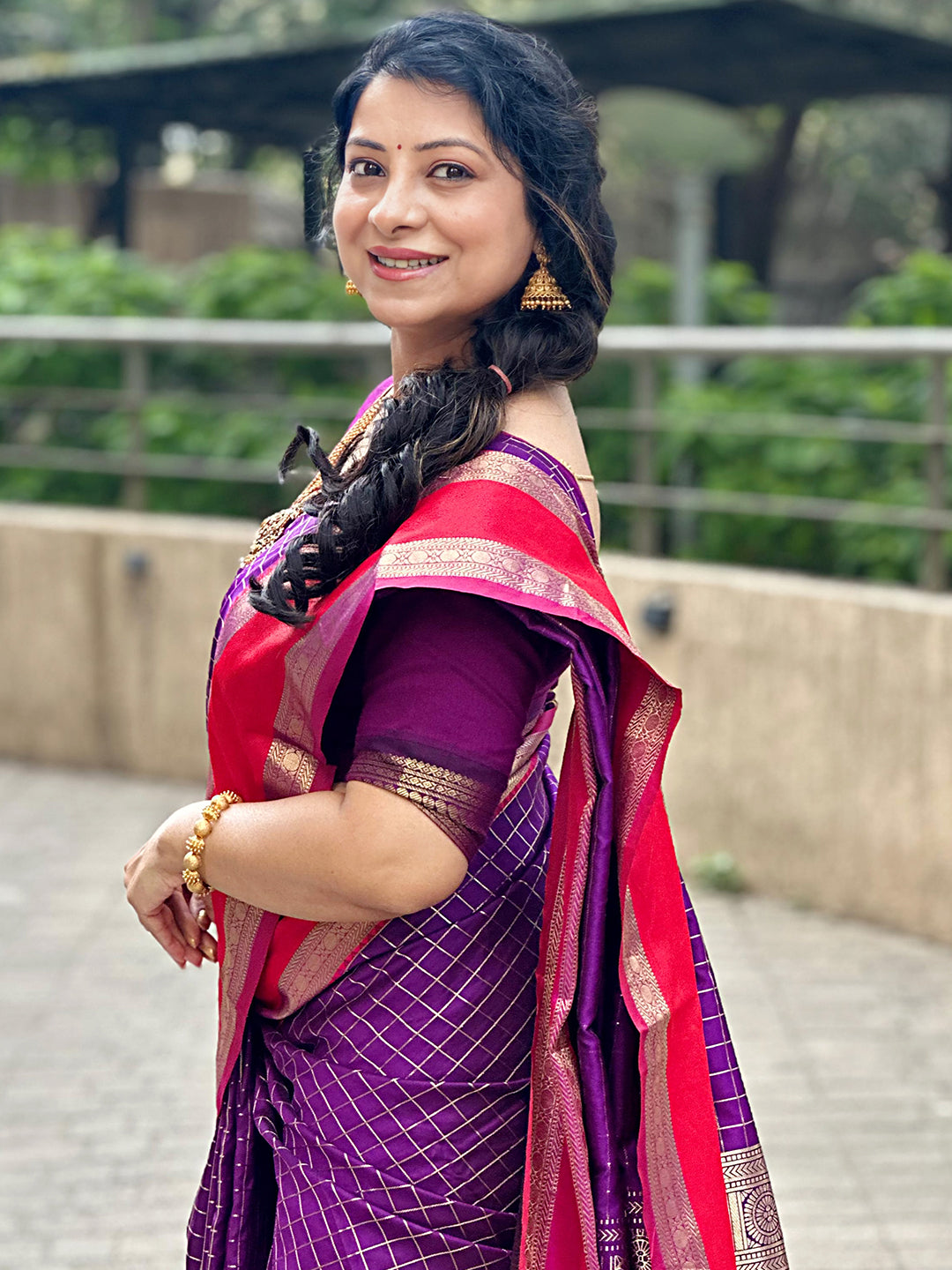 Woman in purple silk saree with red border, smiling sideways.