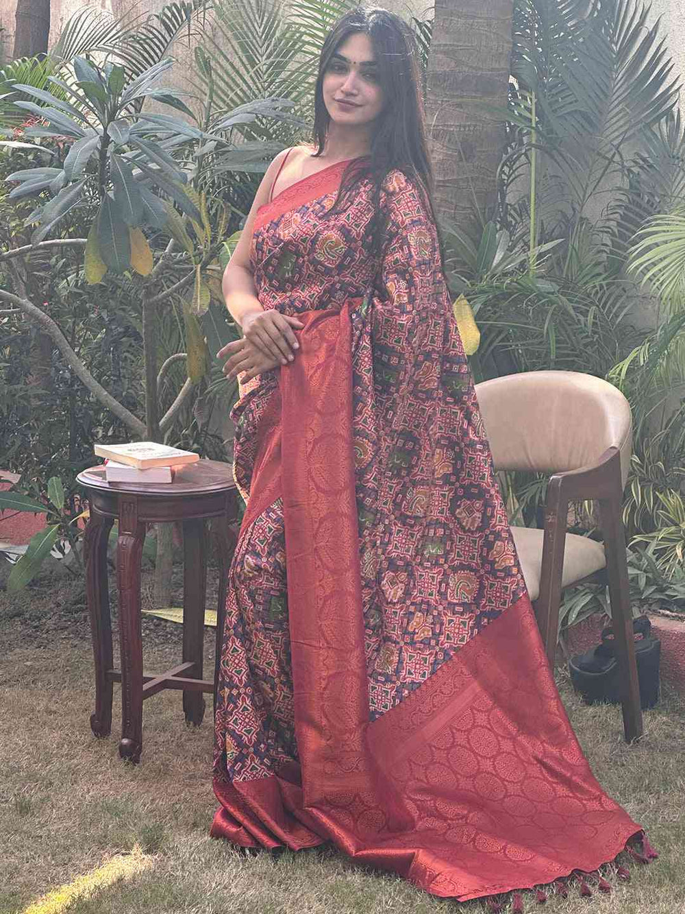 Woman standing in red Patola saree with complex patterns, garden setting.