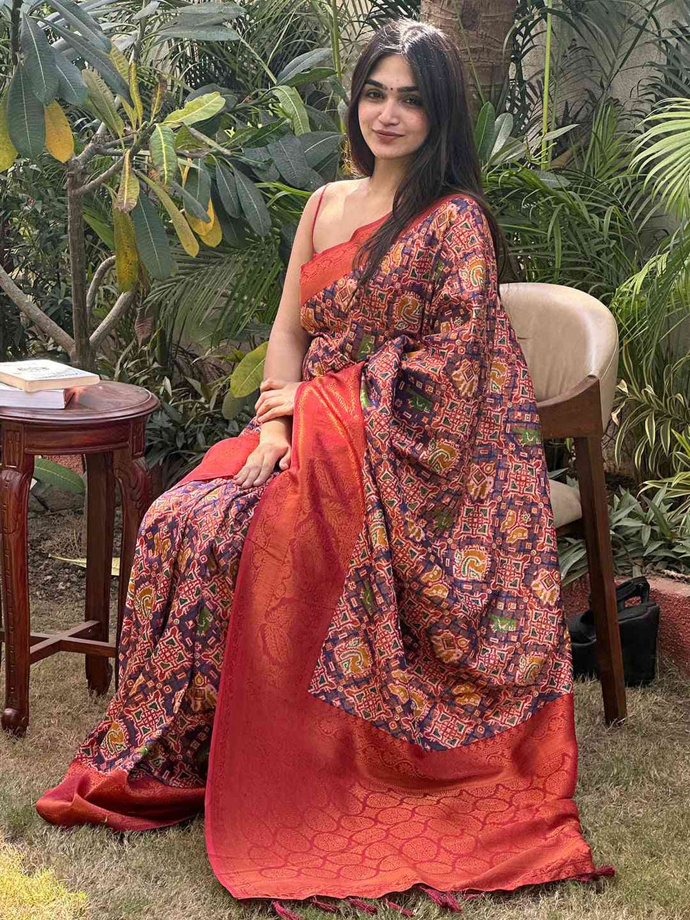 Woman in red Patola saree with intricate patterns, seated outdoors.