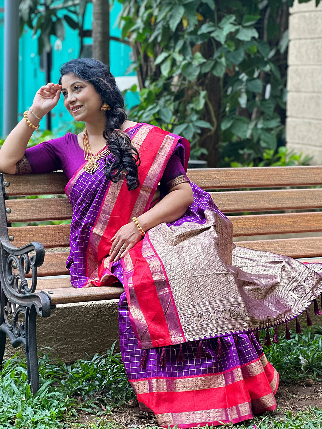 Woman sitting on a bench in purple silk saree with red border.