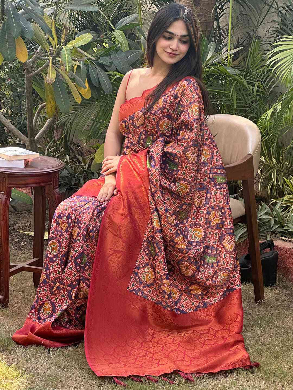 Woman wearing red Patola saree with detailed design, seated outdoors.