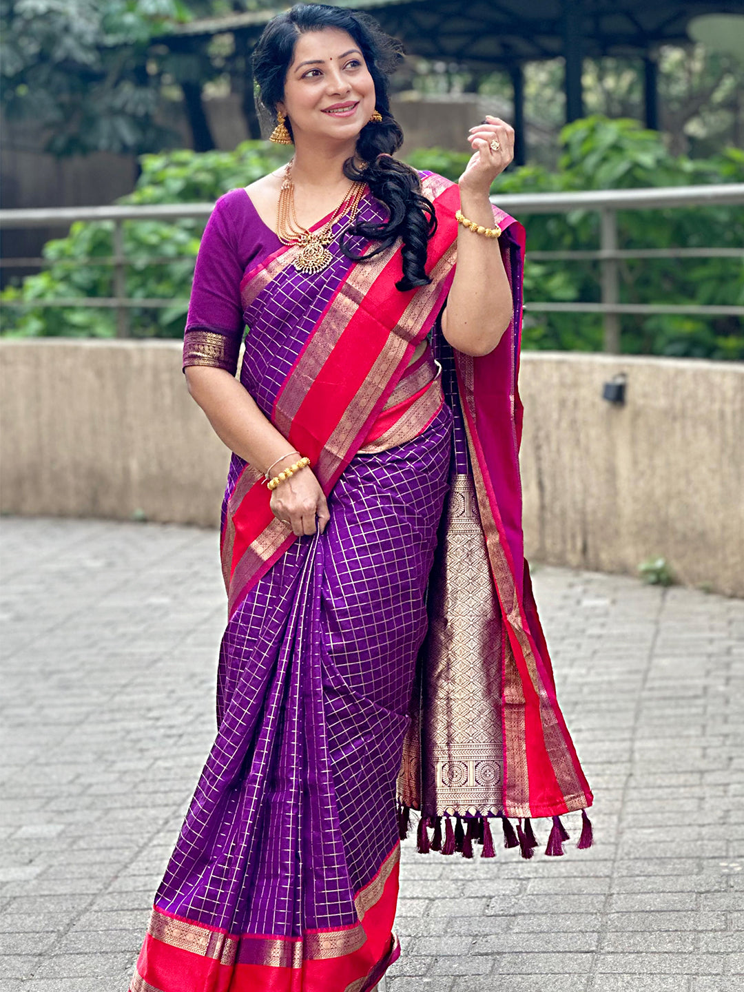 Woman in purple silk saree with red border, posing on a walkway.