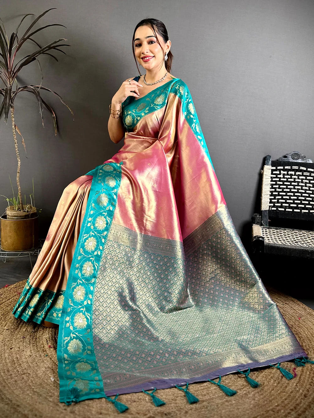 Pink soft tissue zari buti saree, seated pose showing teal woven border and zari pallu with tassels